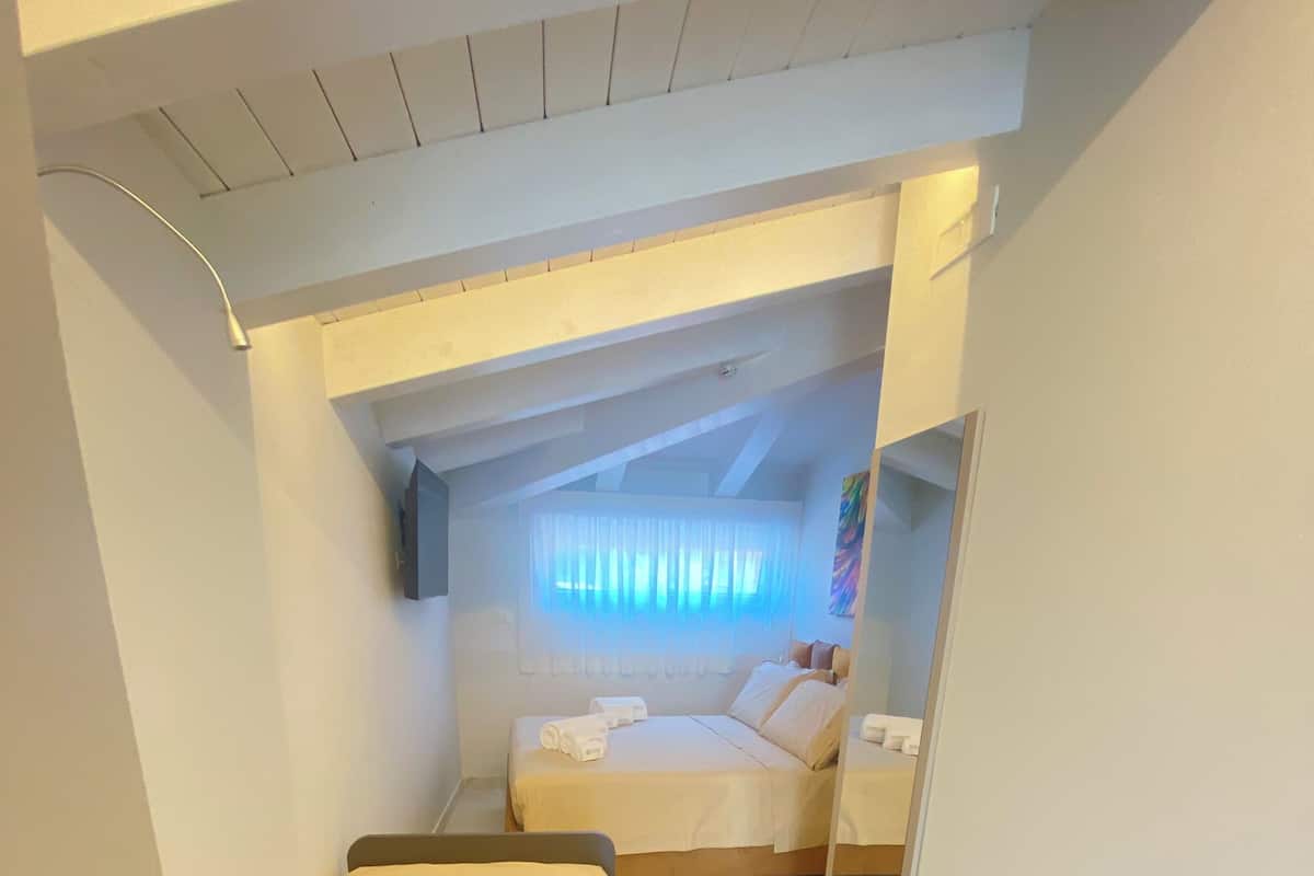 A compact hotel room with a bed, a small bedside table, a mirror, and a window with sheer curtains.