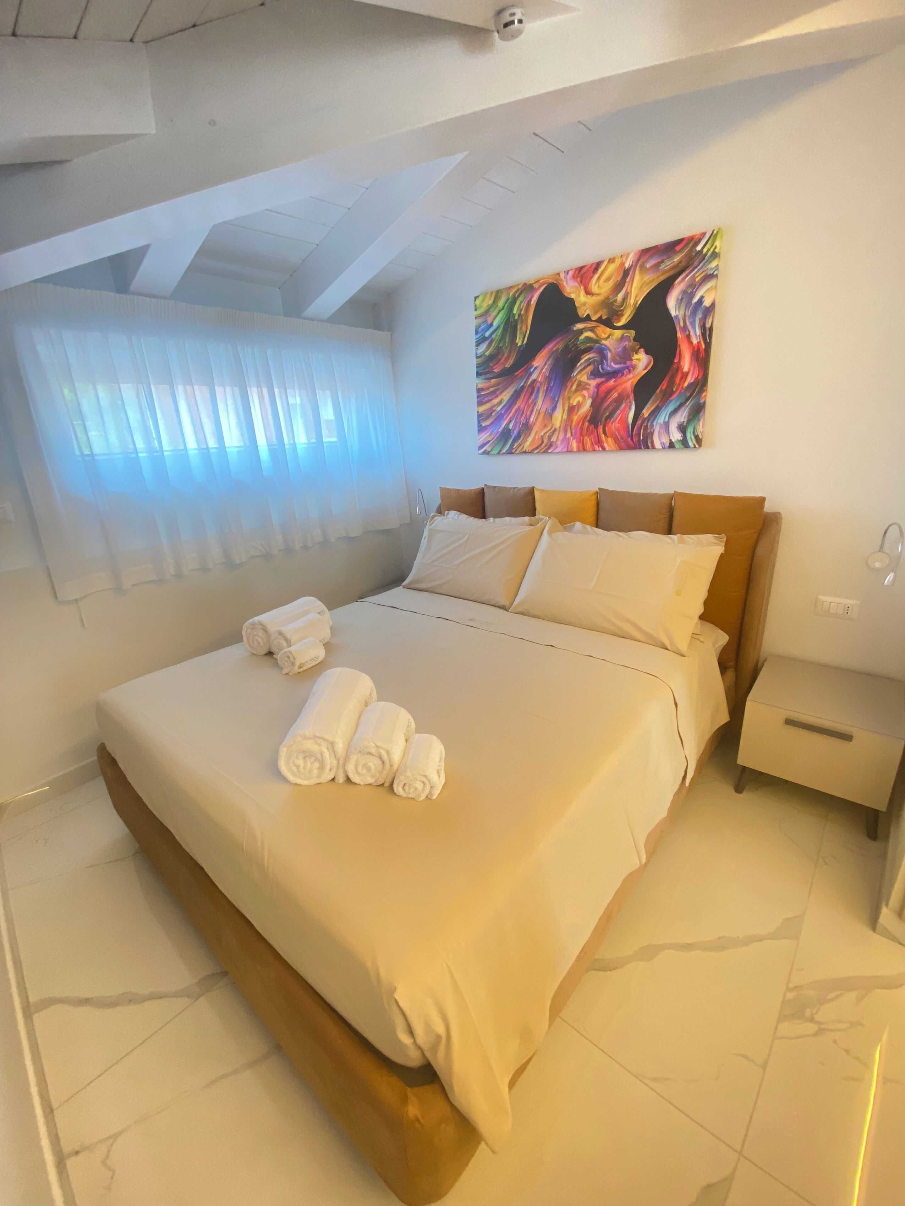 Un dormitorio con una cama, una mesita de noche, un cuadro y una ventana con cortinas.
