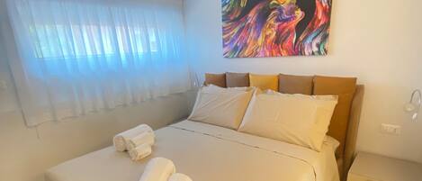 Un dormitorio con una cama, una mesita de noche, un cuadro y una ventana con cortinas.