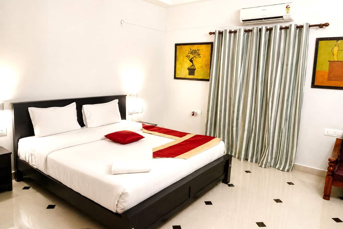 Una habitación de hotel con cama, mesita de noche, equipo de aire acondicionado, ventana con cortinas y obras de arte en las paredes.