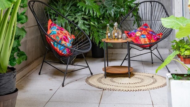 Un jardin intérieur accueillant, agrémenté de plantes suspendues, de deux chaises en fil de fer noir avec des coussins colorés, et d’une petite table en bois sur laquelle sont posées des bouteilles et un objet décoratif.