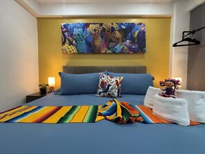 Ein Schlafzimmer mit einem großen Bett, einer farbenfrohen Decke, einem Kissen mit Vogelmotiv, einem Wandbild und einer kleinen Figur auf dem Bett.