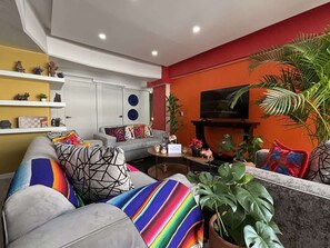 Ein Wohnzimmer mit einer Couch, einem Couchtisch und einem Fernseher. Auf Regalen befinden sich Pflanzen und Dekorationsobjekte.