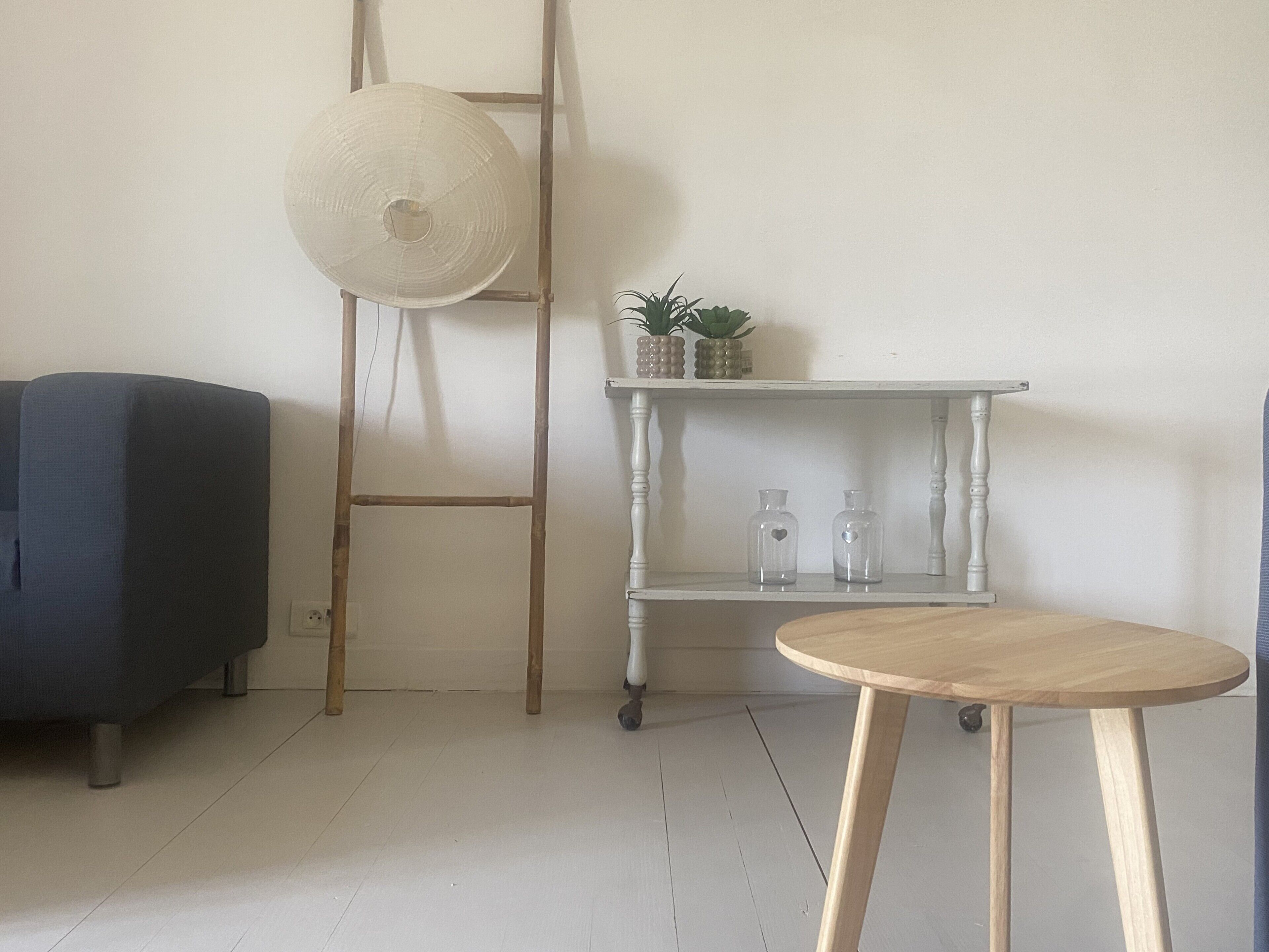 A room with a blue sofa, a wooden ladder, a white shelf with plants and bottles, and a round wooden table.