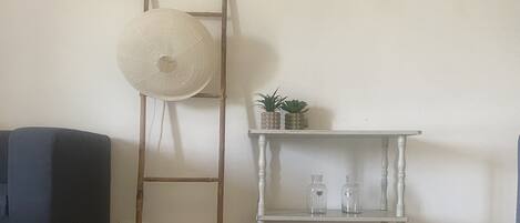 A room with a blue sofa, a wooden ladder, a white shelf with plants and bottles, and a round wooden table.
