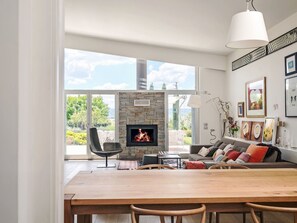 A modern living room with a stone fireplace, a dining area, and large windows offering a view of the outdoors. - Mulberry Garden by Interhome (Belvedere Ostrense)