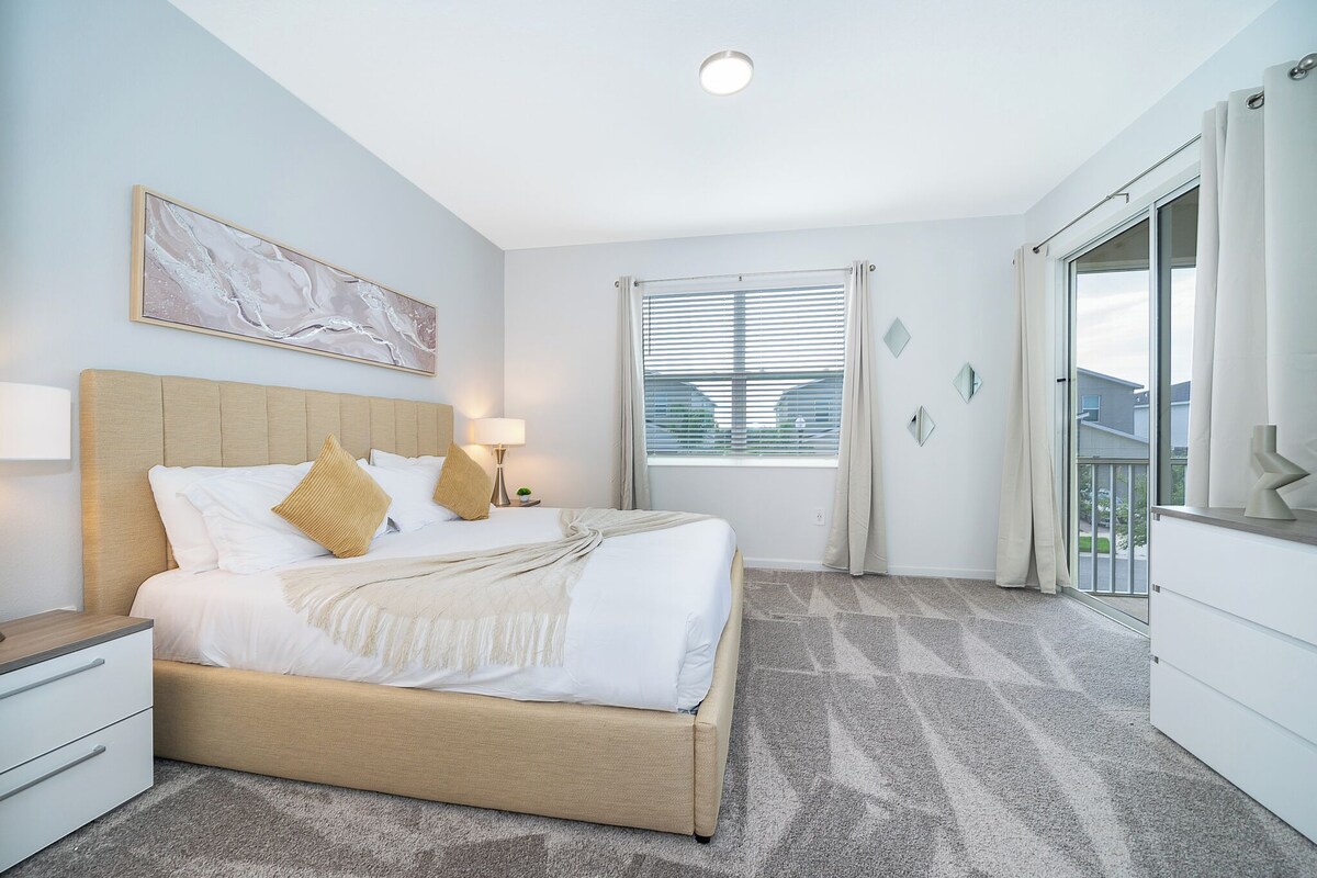 A hotel room with a bed, bedside table, lamp, and a window with blinds.