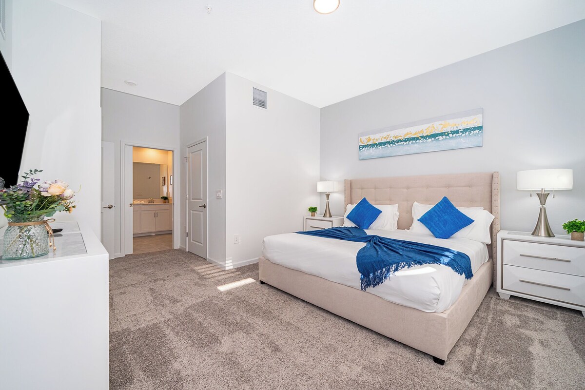 A bedroom with a large bed, bedside tables, a nightstand, a wall-mounted lamp, and a door leading to a bathroom.