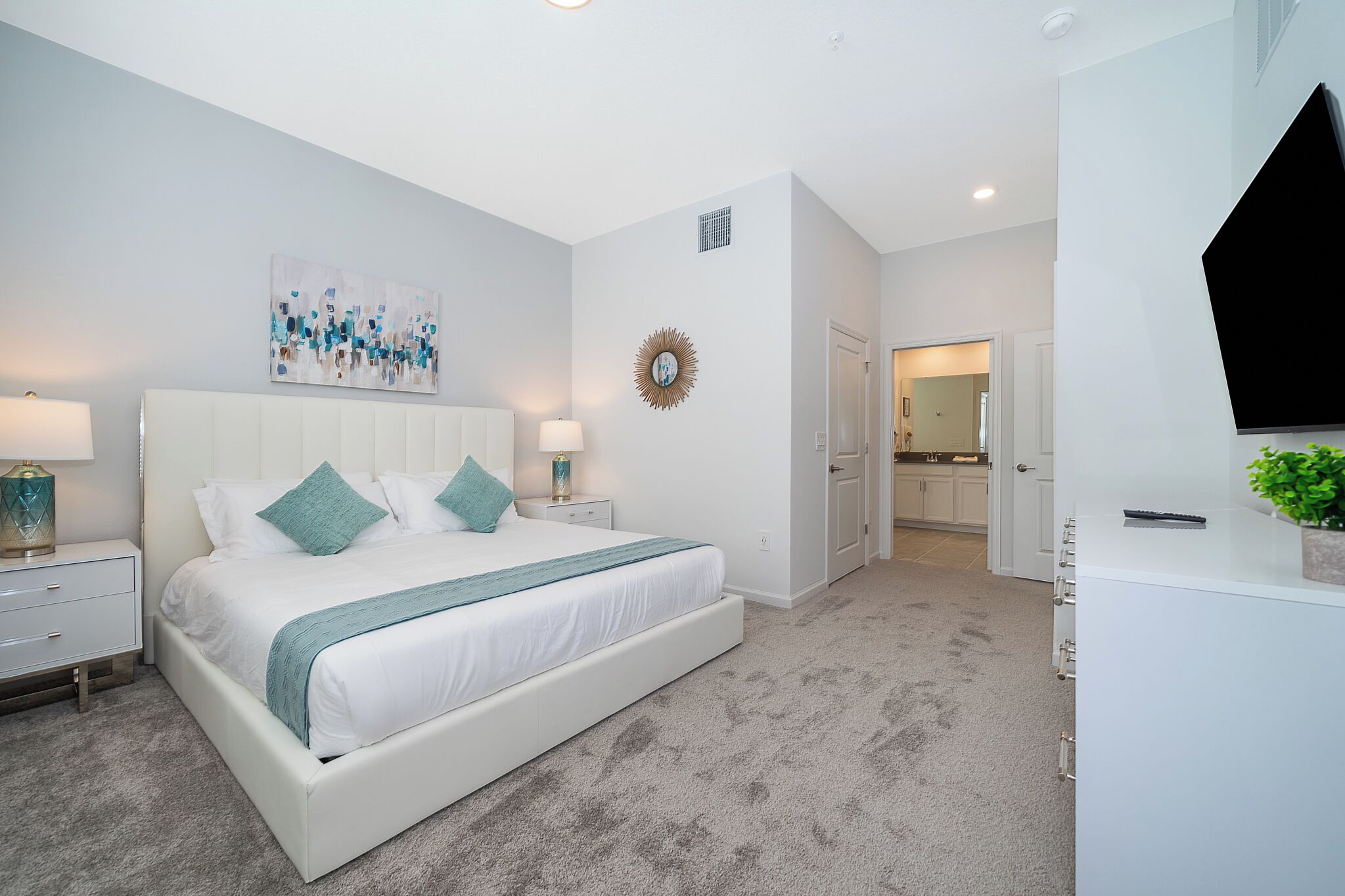 A modern bedroom with a large bed, bedside tables, a TV, and a bathroom visible in the background.
