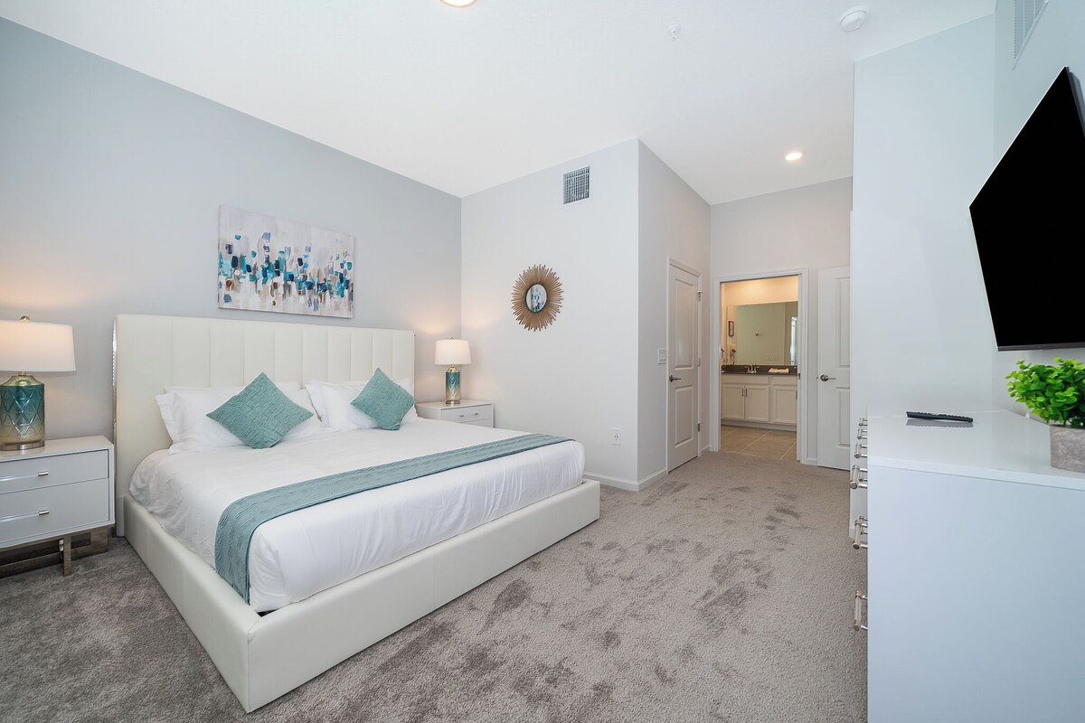 A modern bedroom with a large bed, bedside tables, a TV, and a bathroom visible in the background.