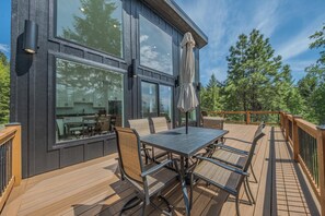 A modern building with a deck featuring a table and chairs, large windows, and a view of trees. - Big Sky Retreat - Mountain Views (Columbia Falls)