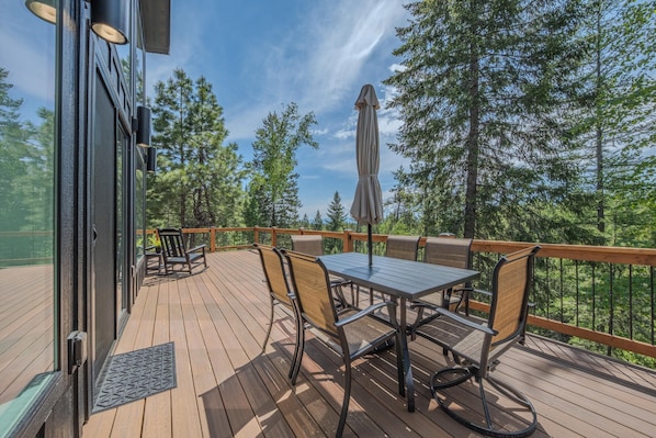 A wooden deck with a table and chairs, surrounded by a railing, overlooking a forested area. - Big Sky Retreat - Mountain Views (Columbia Falls)