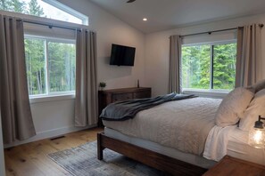 A bedroom with a bed, a TV mounted on the wall, a bedside table, and a window with curtains. - Big Sky Retreat - Mountain Views (Columbia Falls)