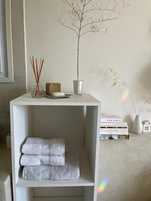 Uma prateleira branca com toalhas dobradas, um vaso com juncos, uma pequena planta em vaso e um quadro na parede.