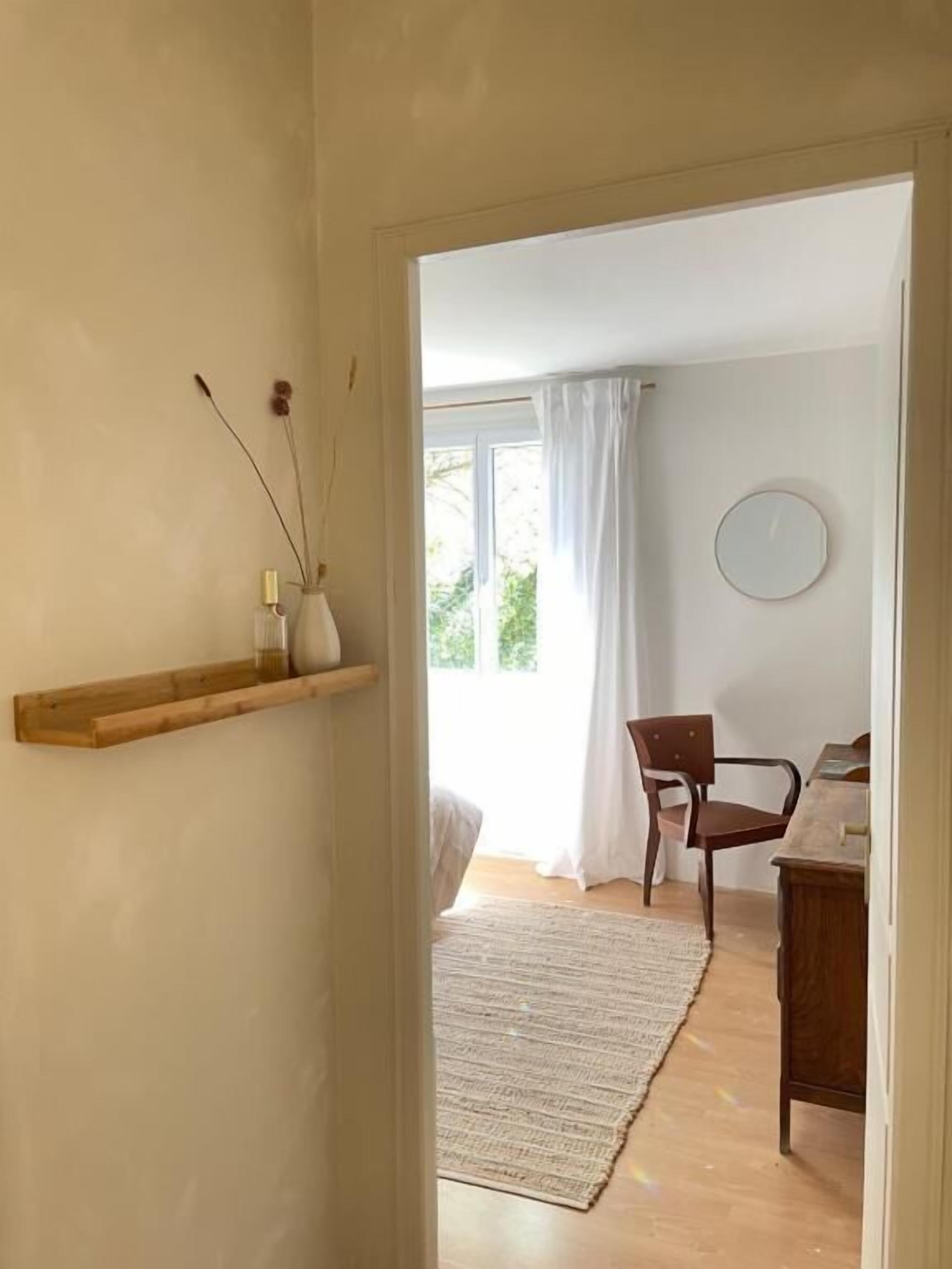 A room with a wooden shelf, a chair, a desk, a rug, and a window with curtains.