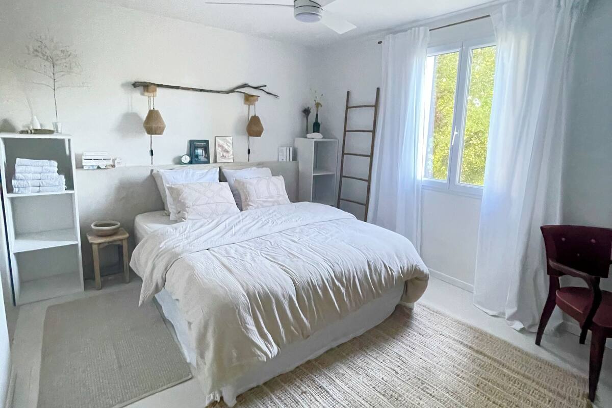 A bedroom with a bed, a ceiling fan, a window with curtains, a bedside table, and a chair.