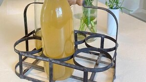 Una botella de líquido amarillo en un soporte metálico, con un jarrón de flores rosadas y una flor roja al fondo.