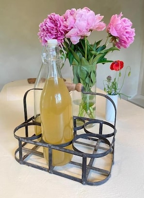 Uma garrafa de líquido amarelo em suporte metálico, com um vaso de flores cor-de-rosa e uma flor vermelha ao fundo.