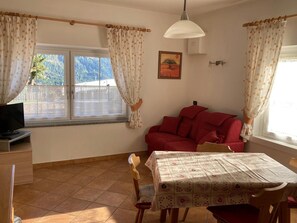 A room with a red sofa, a dining table with a patterned tablecloth, a television on a stand, and a window with curtains. - Apartment 'Cima Dodici' with Mountain View and Wi-Fi (Vigo di Fassa)