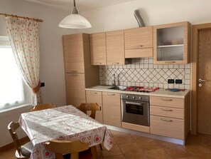 A compact kitchen with a dining table, wooden cabinets, a built-in oven, and a window with curtains. - Apartment 'Cima Dodici' with Mountain View and Wi-Fi (Vigo di Fassa)