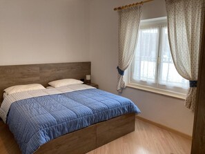 A bedroom with a wooden headboard, a bed with a blue quilt, two pillows, a bedside table with a lamp, and a window with curtains. - Apartment 'Cima Dodici' with Mountain View and Wi-Fi (Vigo di Fassa)