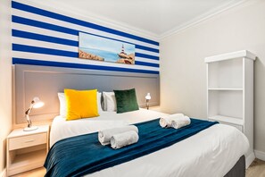 A hotel room with a bed, pillows, a nightstand, a lamp, a painting, and a white shelf. - Ocean View C303 (Cape Town)