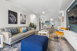 A modern living room with a sofa, ottoman, and coffee table, a kitchen in the background, and artwork on the walls. - Ocean View C303 (Cape Town)