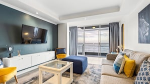 A modern living room with a flat-screen TV, a sofa, a coffee table, and a view of the sea through large windows. - Ocean View C303 (Cape Town)
