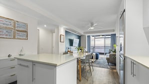 A modern kitchen with a central island, a dining area with chairs, a sofa, and a view of the outdoors. - Ocean View C303 (Cape Town)