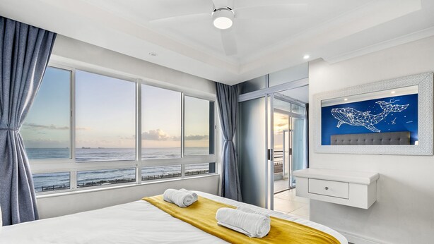 A hotel room with a large window overlooking the sea, a bed with white linens, a yellow throw blanket, and a wall-mounted artwork. - Ocean View C303 (Cape Town)
