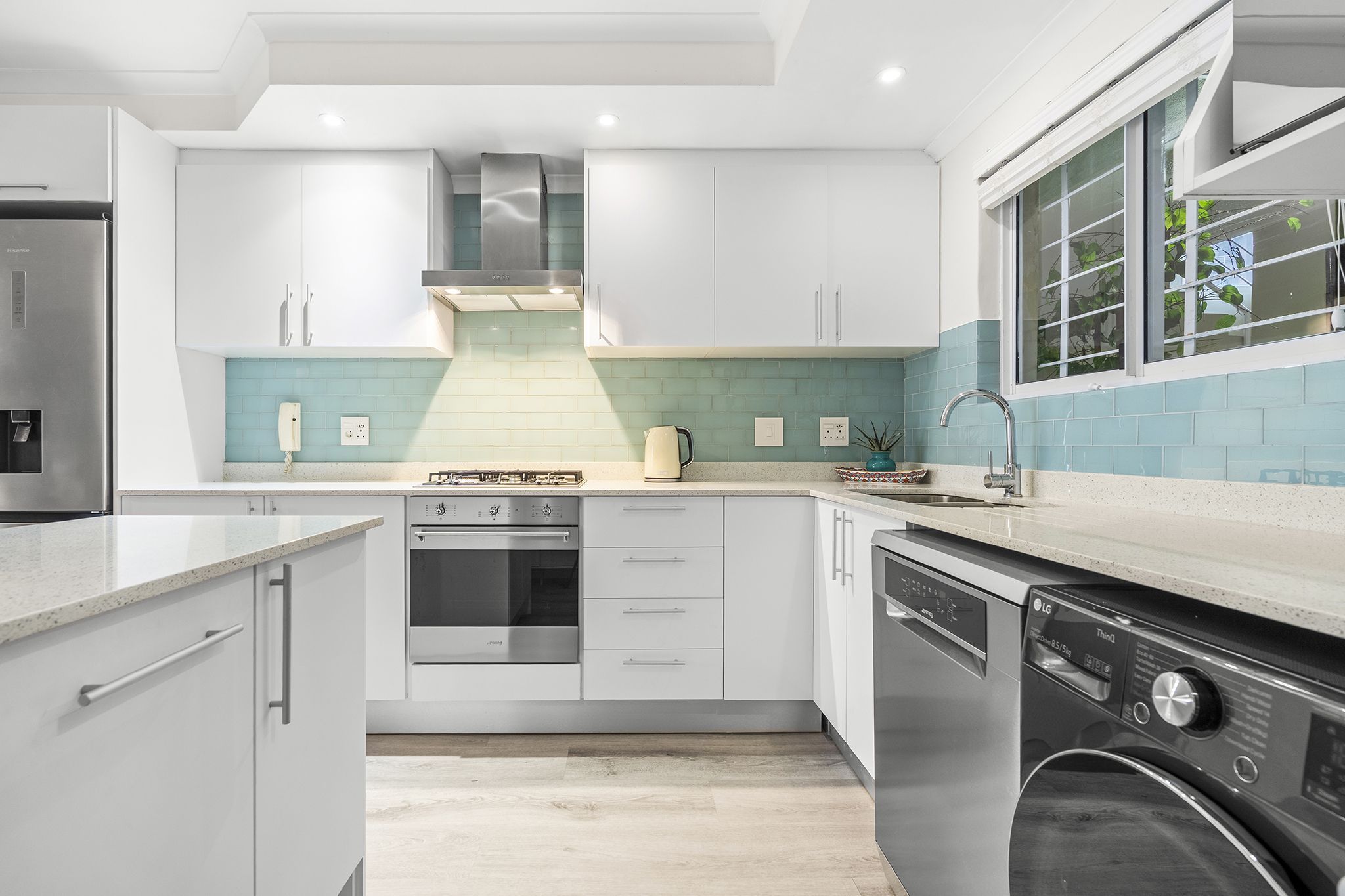 A modern kitchen with white cabinets, a stainless steel range hood, a built-in oven, a dishwasher, and a window with a view of greenery.