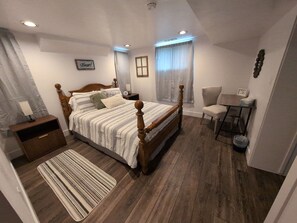 Een slaapkamer met een houten baldakijnbed, een nachtkastje met een lamp, een stoel, een klein tafeltje met een fotolijst, en een raam met gordijnen.