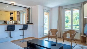 A modern living room with a sofa, two chairs, a coffee table, and a TV. There is a kitchen area with a counter and a mirror. - Les Balmettes I T3 I Annecy (Annecy)