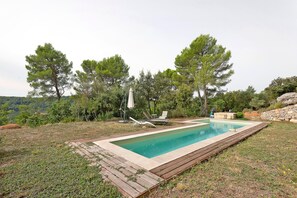 Una piscina rectangular con una terraza de madera alrededor, dos sillas de descanso y un muro de piedra al fondo.