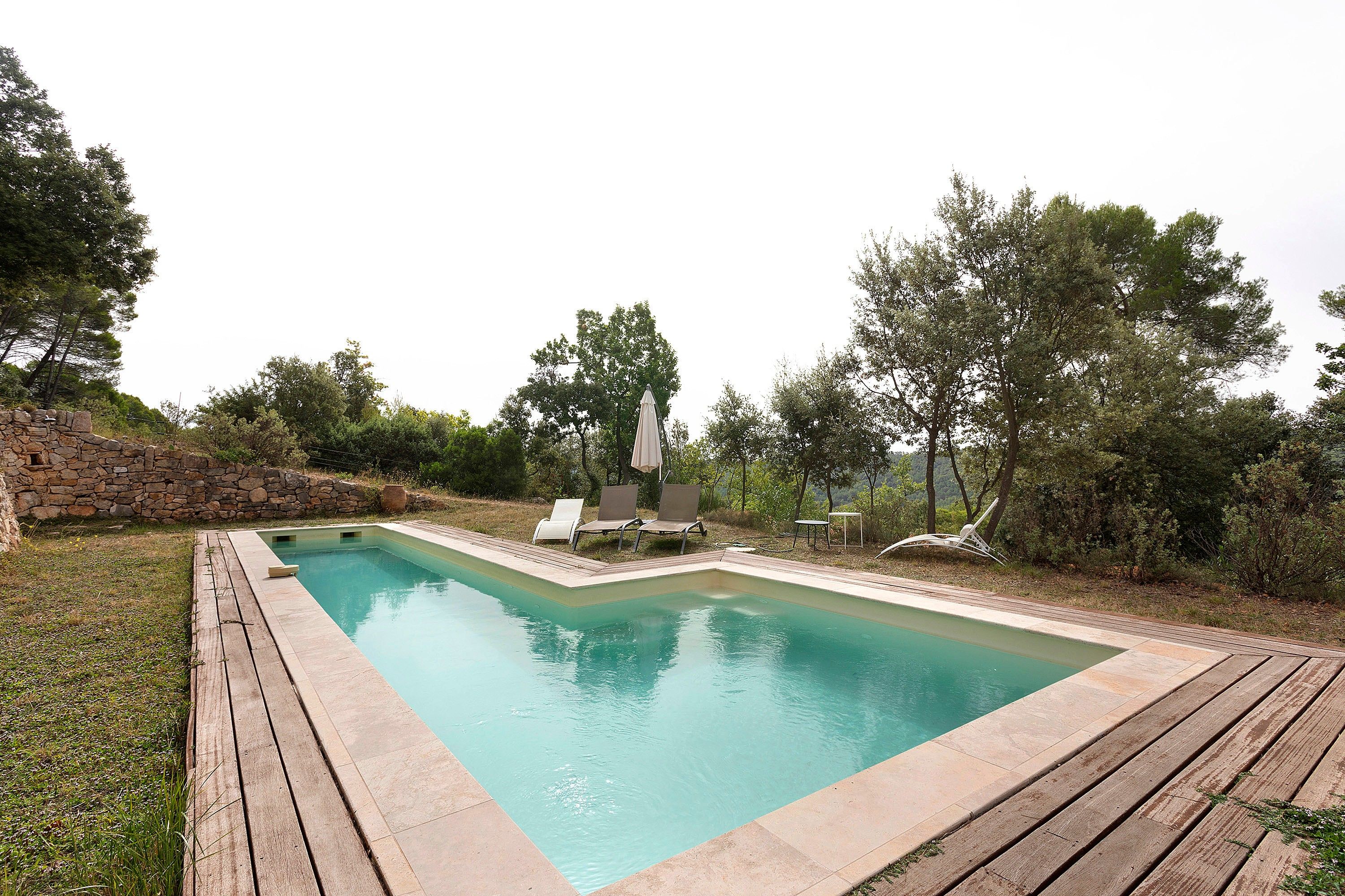 Ein rechteckiger Pool mit einer Holzterrasse darum, einer Steinmauer an einer Seite und einigen Liegestühlen und Sonnenschirmen im Hintergrund.