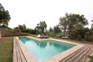 Una piscina rectangular con una cubierta de madera que la rodea, un muro de piedra en un lateral y algunas tumbonas y sombrillas al fondo.