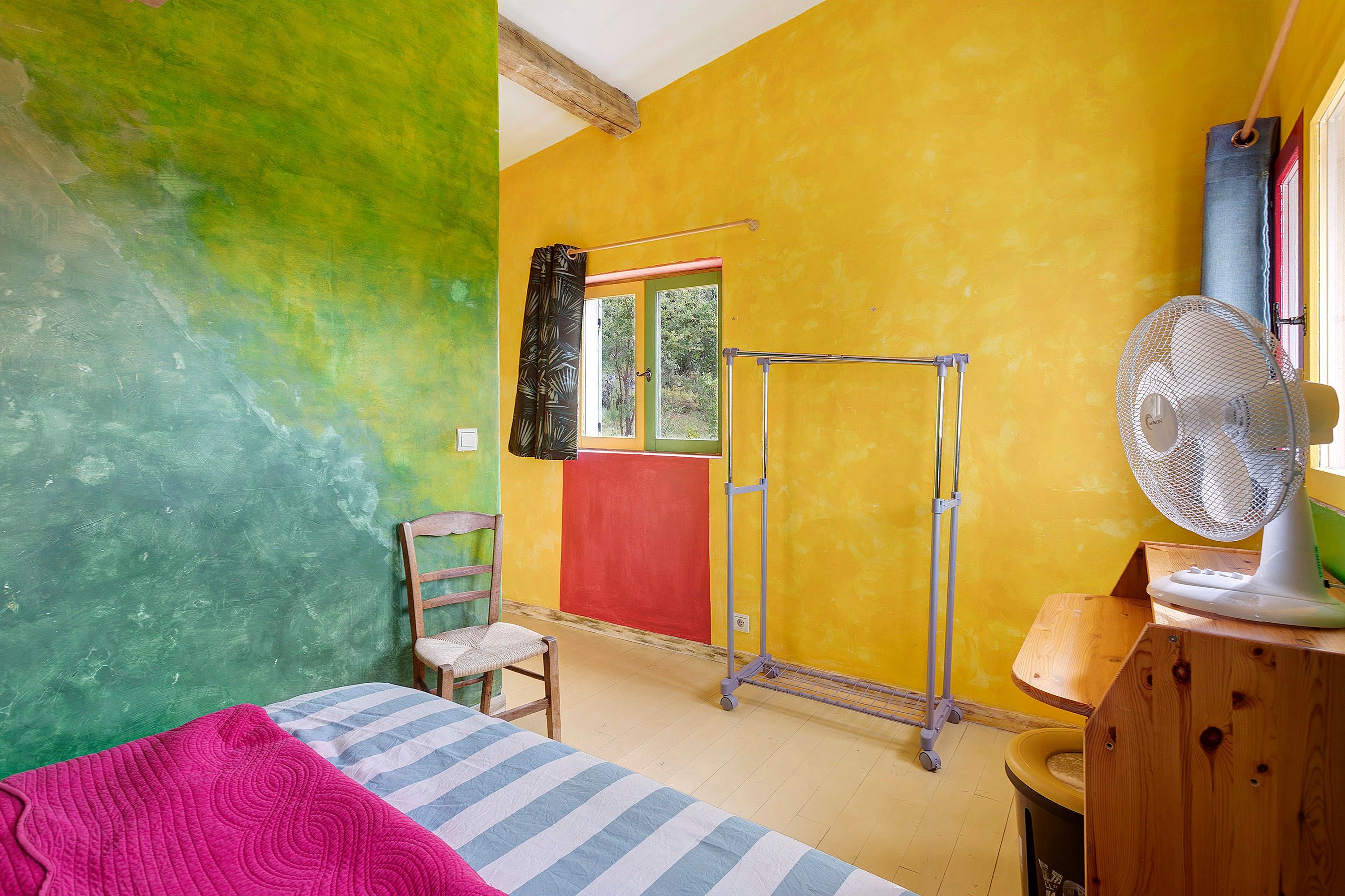 Ein Zimmer mit einem Bett, einem Sessel, einem Fenster mit Blick auf Bäume, einem Ventilator und einer Tür.