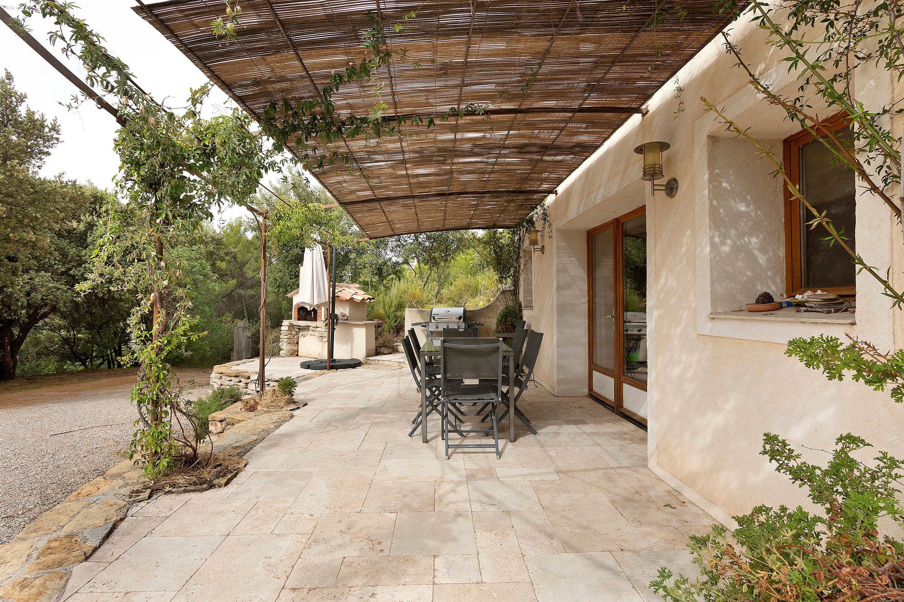 Ein Außenbereich mit Terrasse, einem Pergola-Überdachung, Steinböden und einem Esstisch mit Stühlen.