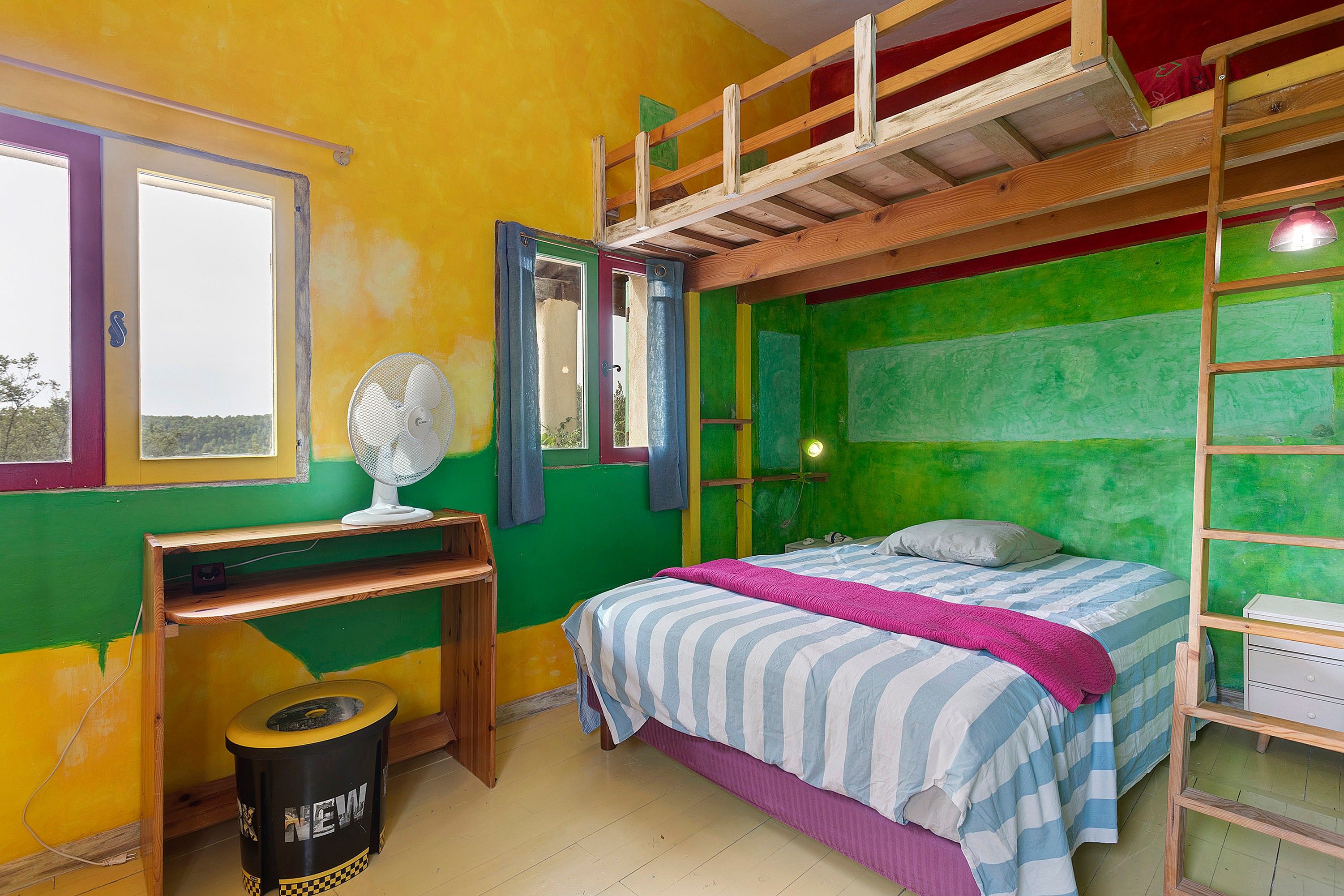 A room with a bunk bed, a desk with a chair, a standing fan, and a small trash bin.