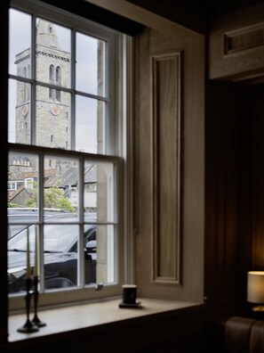 Ein Zimmer mit großem Fenster, im Blick ein Glockenturm, ein Tisch mit Kerze und eine Lampe.