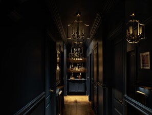 Ein schmaler Korridor mit dunklen Wänden, einem Holzboden und einer zentralen Chandelier. Regale mit Flaschen und Gläsern säumen die Wände.