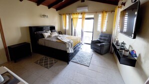 A bedroom with a bed, a chair, a TV, and a window with curtains. - Haversville Canary Studio with stunning ocean views on Tortola, Virgin Islands (Tortola)