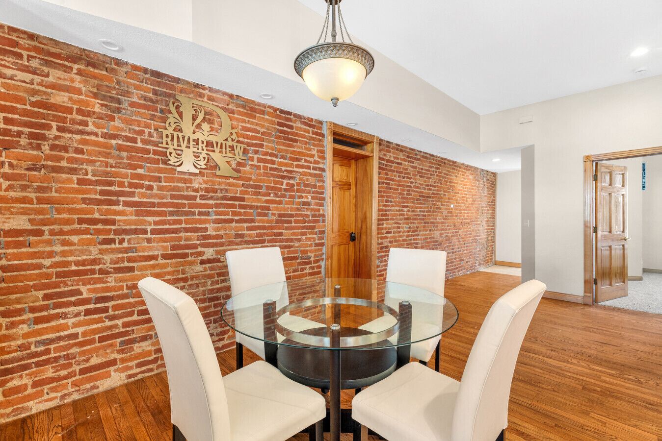 Una habitación con pared de ladrillo, una mesa redonda de cristal con cuatro sillas, una puerta de madera y una lámpara colgante.