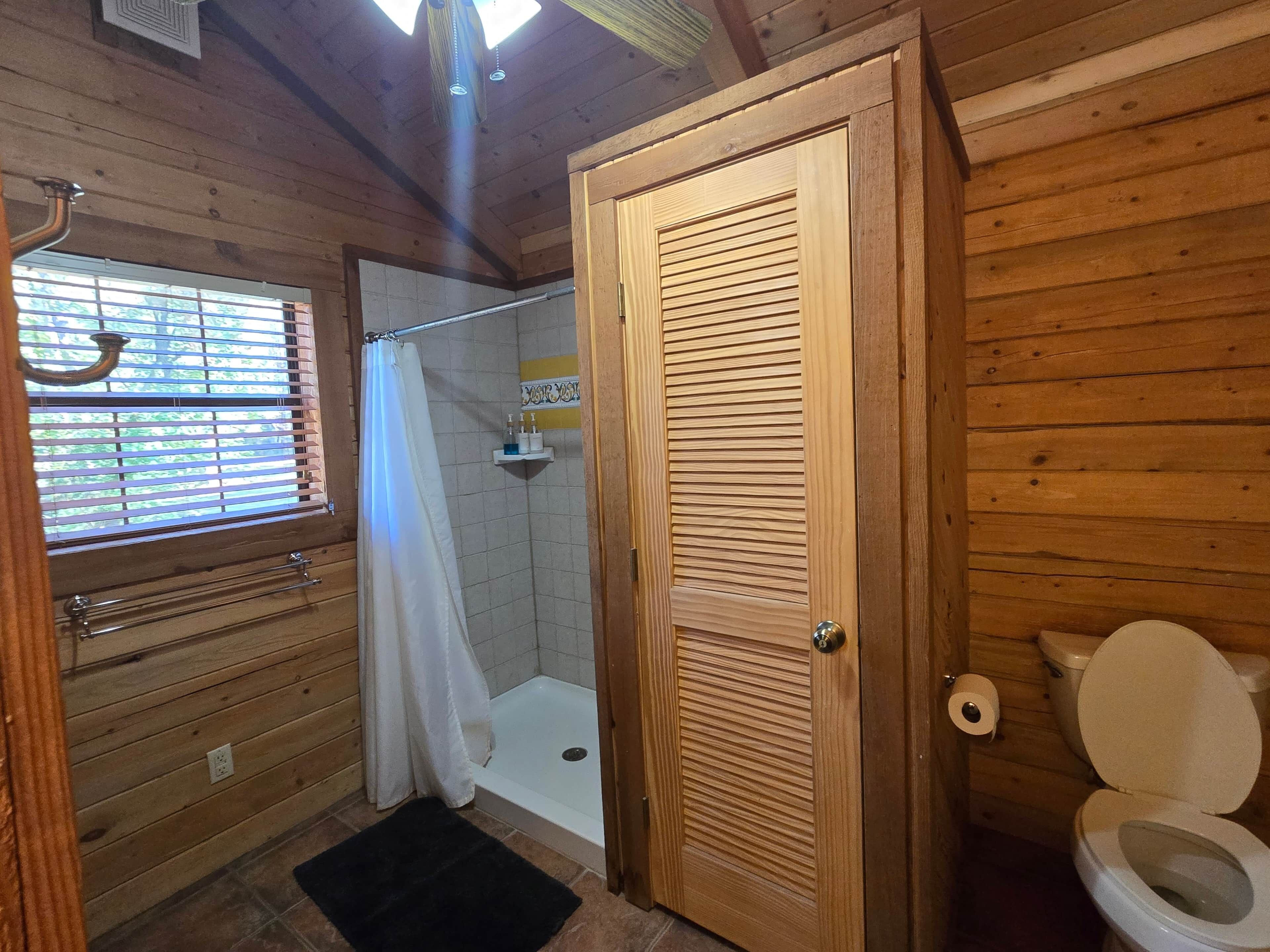 A wooden cabin bathroom with a shower, toilet, and window with blinds.