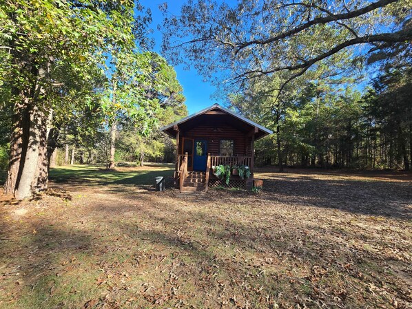 A rustic cabin with a porch, surrounded by trees and a grassy area.