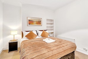 A neatly arranged bedroom with a bed, a bedside lamp, a painting on the wall, and a radiator.