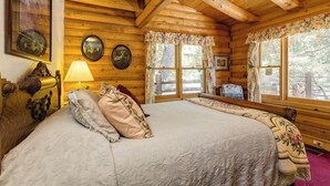Una habitación de cabaña de madera con una cama, dos ventanas con cortinas, una lámpara y cuadros colgados en la pared.