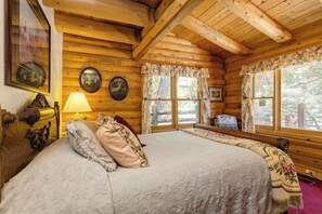A wooden cabin room with a bed, two windows with curtains, a lamp, and wall-mounted pictures. - This newer, spacious 2600 sq. ft 2-story log home is well insulated with thermostatically controlled central heating.  There are 4 large bedrooms and the Master Suite is complete with a king size brass bed with a separate sitting area, 25" Color Tele (Idyllwild Ca 92549)