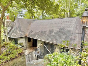 A rustic stone house with a steeply pitched roof, a small porch with a metal railing, and a lantern-style light fixture.