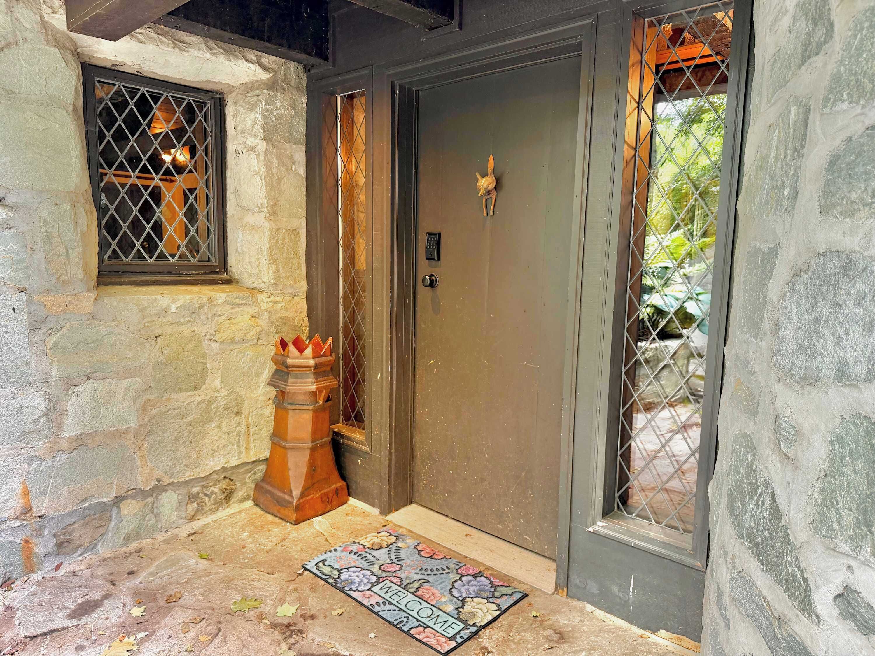 Una entrada de paredes de piedra con una puerta oscura, un pequeño ventanal con reja de metal, un objeto decorativo de madera y un felpudo.